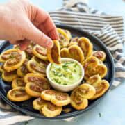 Hand dipping a flaky pesto and cheese palmier into a creamy herb sauce, highlighting the crisp texture and soft, cheesy center.