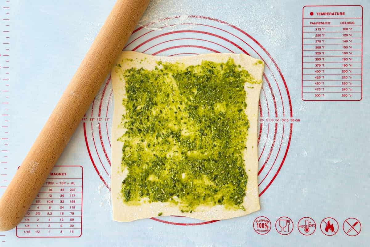 Puff pastry rolled into a square and spread evenly with vibrant green basil pesto on a lightly floured silicone baking mat.