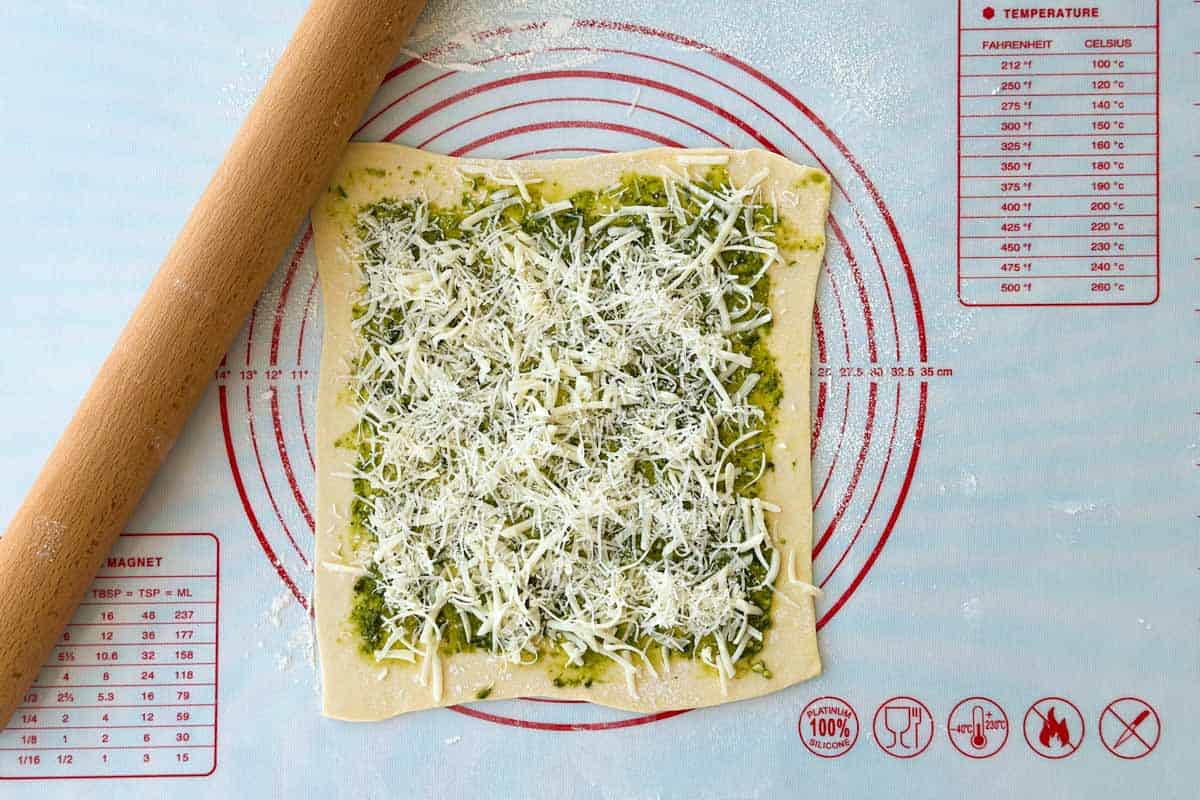 Puff pastry topped with basil pesto and generously sprinkled with grated cheese, ready for shaping into savory palmiers.
