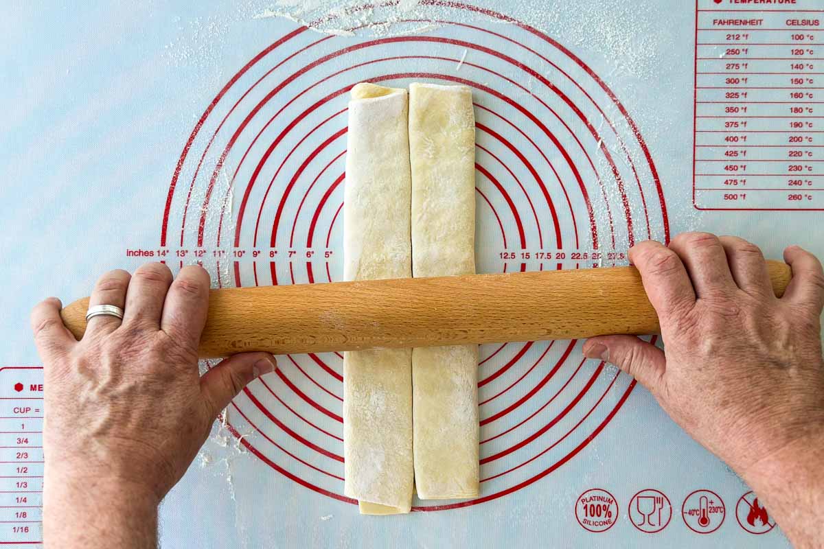 Hands using a rolling pin to gently press and flatten the folded puff pastry log for even shaping.