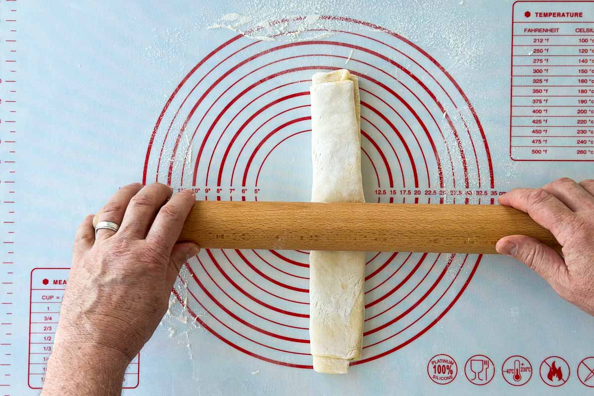 Rolled puff pastry log being flattened further with a rolling pin to create a compact and even thickness.