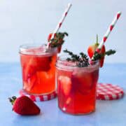 Two strawberry gin and tonic cocktails in small jars, filled with ice and sliced strawberries, garnished with thyme and paper straws.