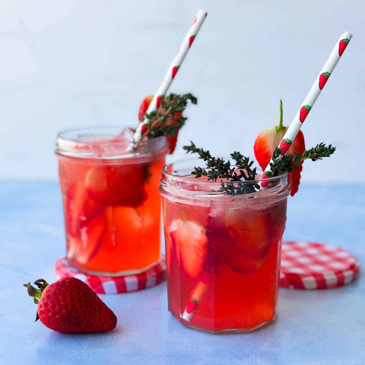 Two strawberry gin and tonic cocktails in small jars, filled with ice and sliced strawberries, garnished with thyme and paper straws.