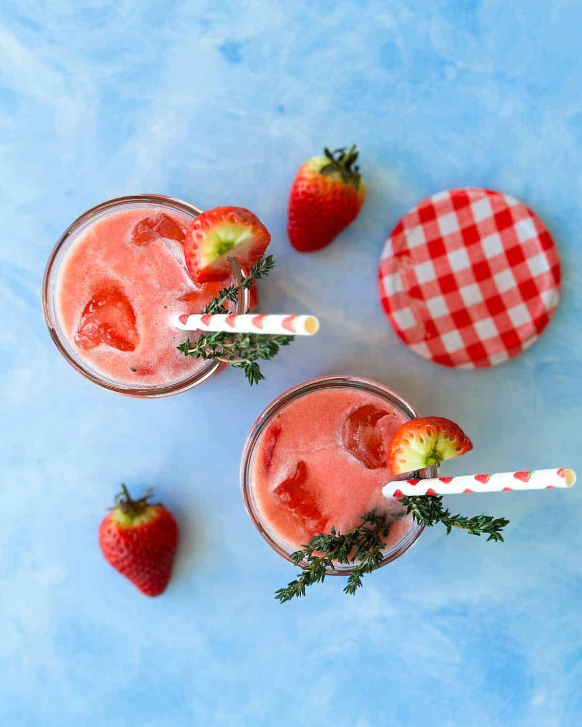 Strawberry gin and tonic cocktails photographed from above with fresh strawberries and gingham jar lids on a blue surface.
