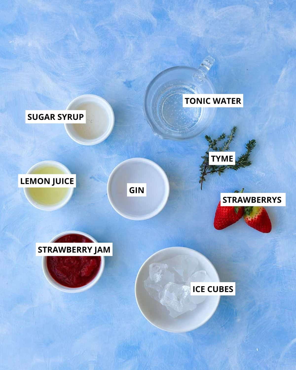 Strawberry gin and tonic ingredients laid out on a blue background including gin, tonic water, strawberry jam, fresh strawberries, lemon juice, sugar syrup, ice cubes, and fresh thyme.