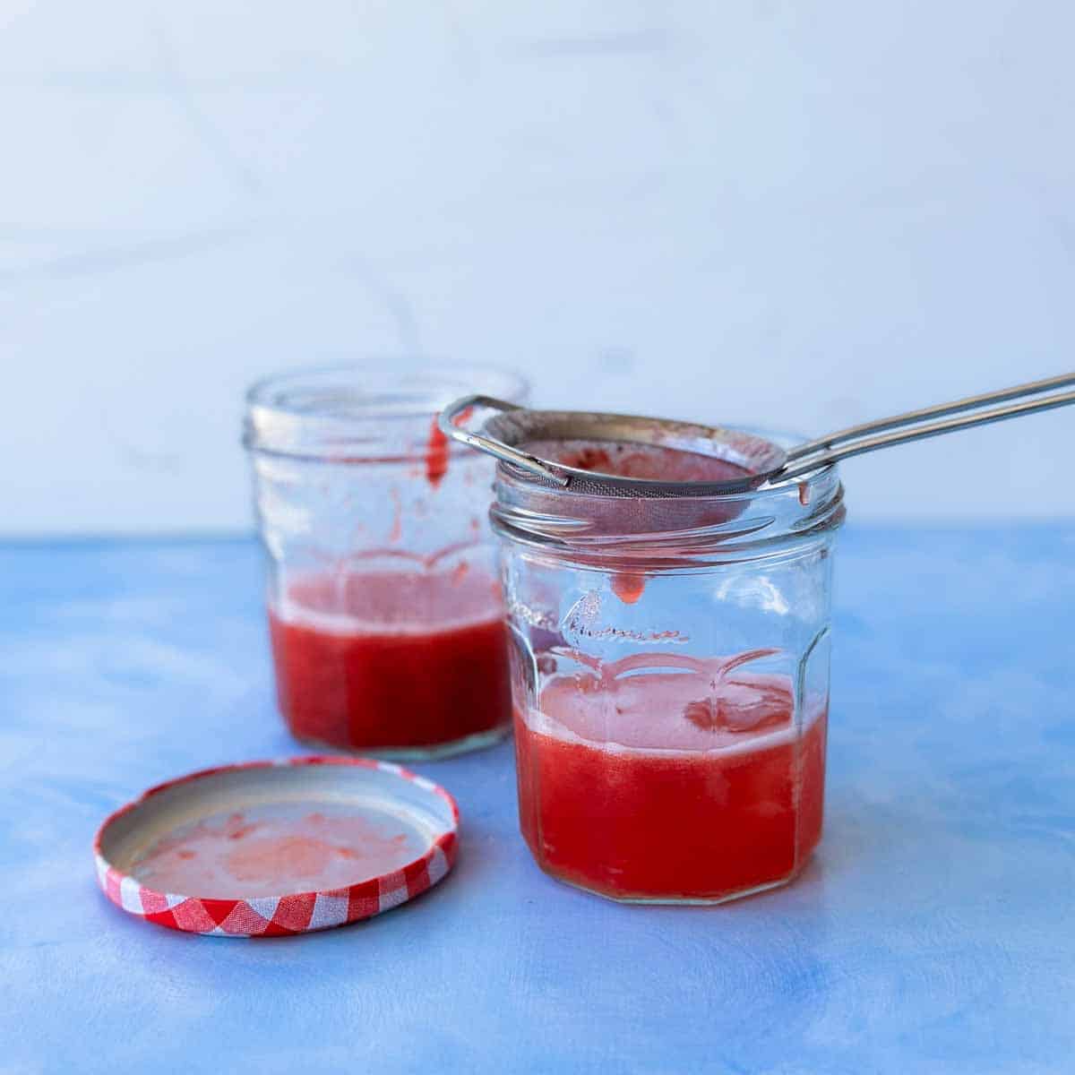 Straining the strawberry jam, gin, lemon juice and sugar syrup mixture through a fine mesh sieve into two glass jars, with ice cubes, before topping up with tonic and garnishing .