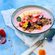 Side angle shot of a thick strawberry smoothie bowl garnished with banana slices, fresh strawberries, granola, mint, and edible flowers, served in a ceramic bowl with a spoon on a blue surface.