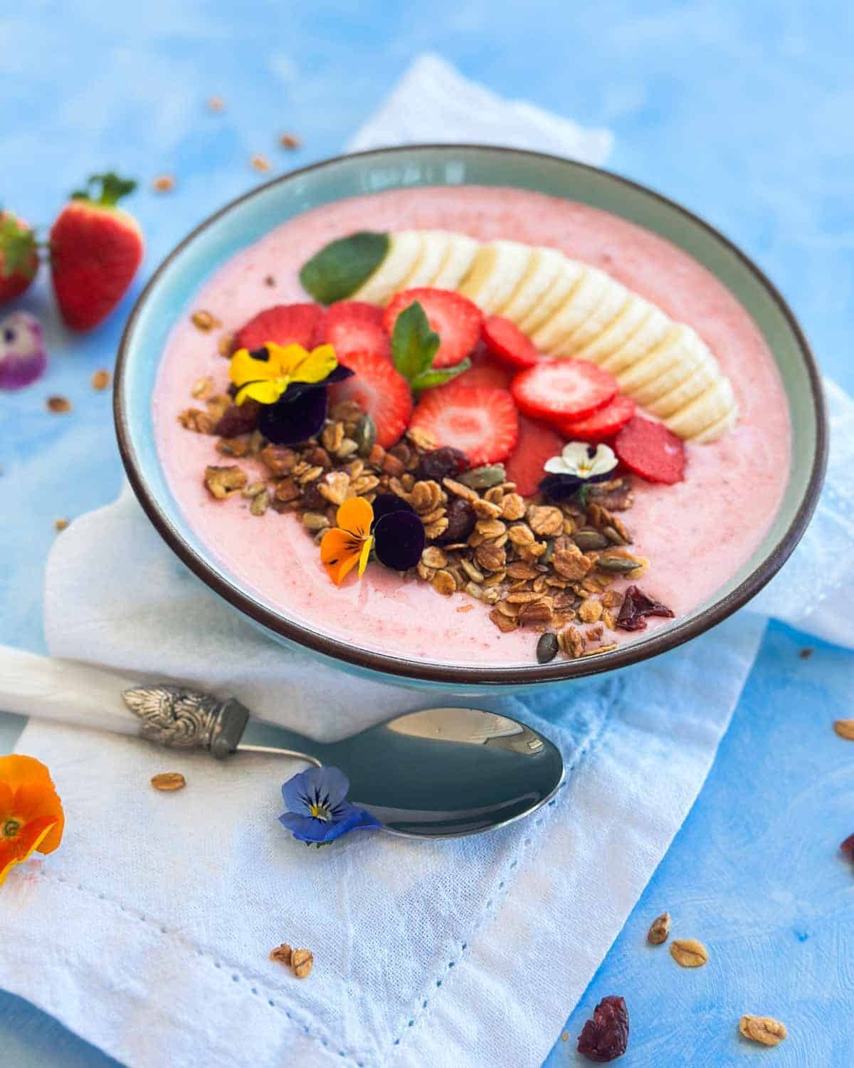 Angled close-up of a strawberry banana smoothie bowl with crunchy granola and edible flowers, styled with a silver-handled spoon and fresh strawberries on a light blue background.
