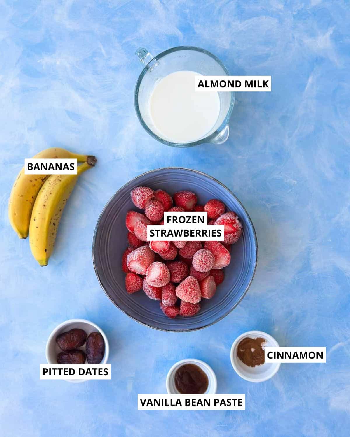 Overhead image of the strawberry smoothie bowl ingredients including frozen strawberries, ripe bananas, almond milk, pitted dates, vanilla bean paste, and ground cinnamon arranged on a light blue surface with clear labels.
