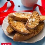 Plate of easy homemade almond biscotti slices with whole almonds, styled with red ribbon and coffee cups. Crisp golden texture highlighted for a simple Italian cookie perfect for gifting or dipping.