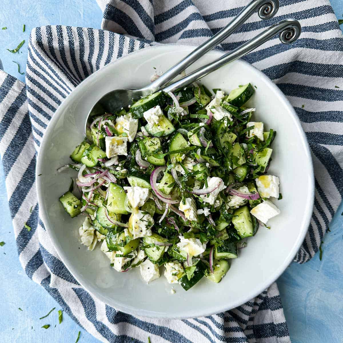Cucumber and feta salad with red onion and fresh herbs served in a white bowl with two spoons on a striped cloth over a light blue background.