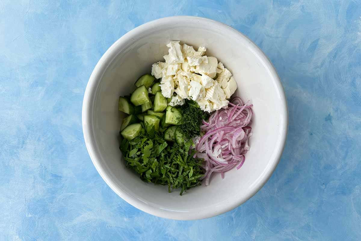 Chopped cucumbers, sliced red onion, crumbled feta, and fresh herbs in a white bowl ready to be mixed for cucumber and feta salad.