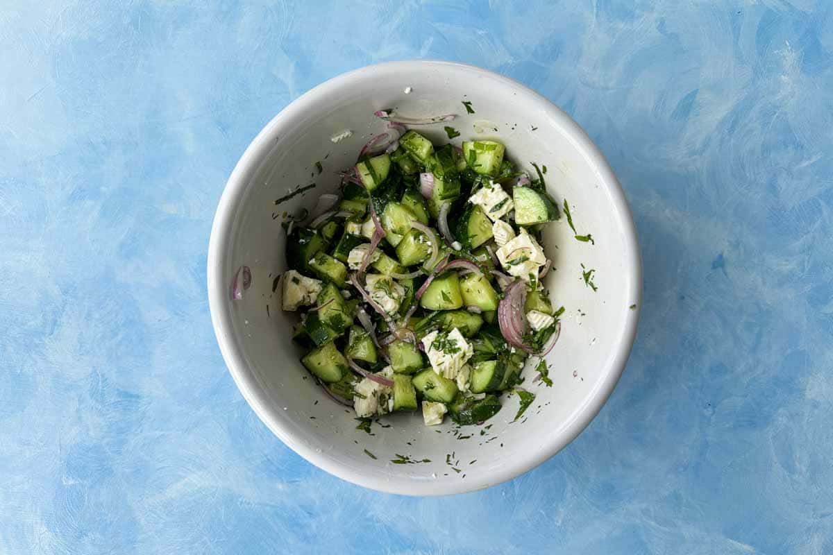 Cucumber and feta salad mixed together in a white bowl with chopped cucumbers, fresh herbs, red onion, and crumbled feta.