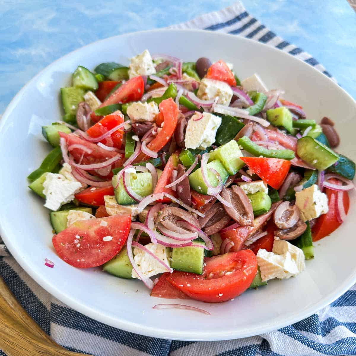 Close-up angled view of Greek salad showing juicy tomato wedges, crisp cucumber, thinly sliced red onion, olives and creamy feta, lightly dressed and glistening in a white serving bowl.