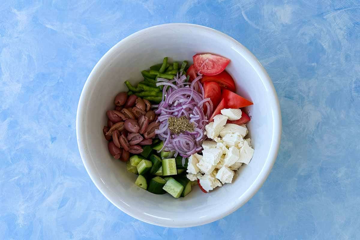 Top-down view of a white mixing bowl filled with chopped cucumber, tomato wedges, sliced red onion, olives, feta and oregano before mixing, arranged neatly for preparation.