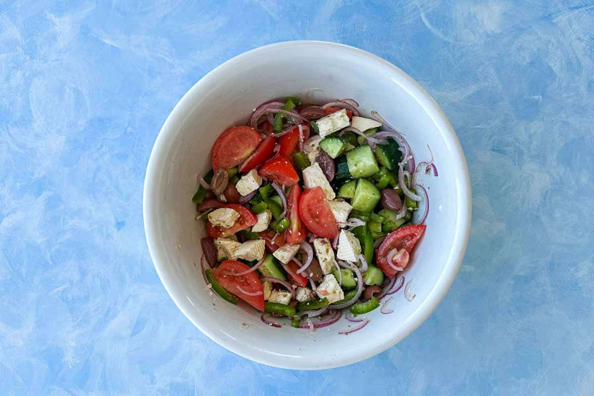 Overhead view of Greek salad being mixed in a white bowl, showing the ingredients lightly combined with dressing, creating a colorful and fresh Traditional Greek salad.