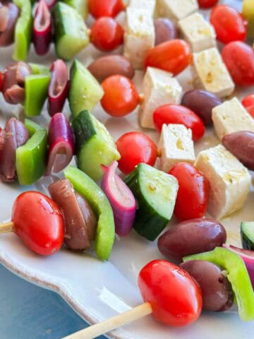 Close-up of Greek salad skewers with cherry tomatoes, Kalamata olives, cucumber, green bell pepper and pickled red onion alongside feta cheese.