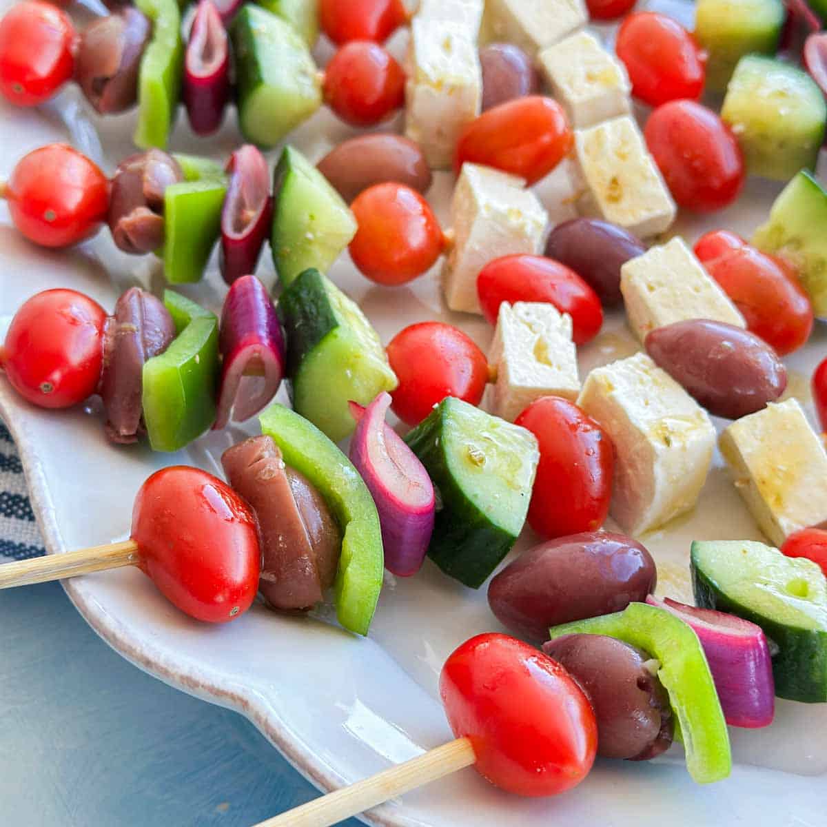 Close-up of Greek salad skewers with cherry tomatoes, Kalamata olives, cucumber, green bell pepper and pickled red onion alongside feta cheese.