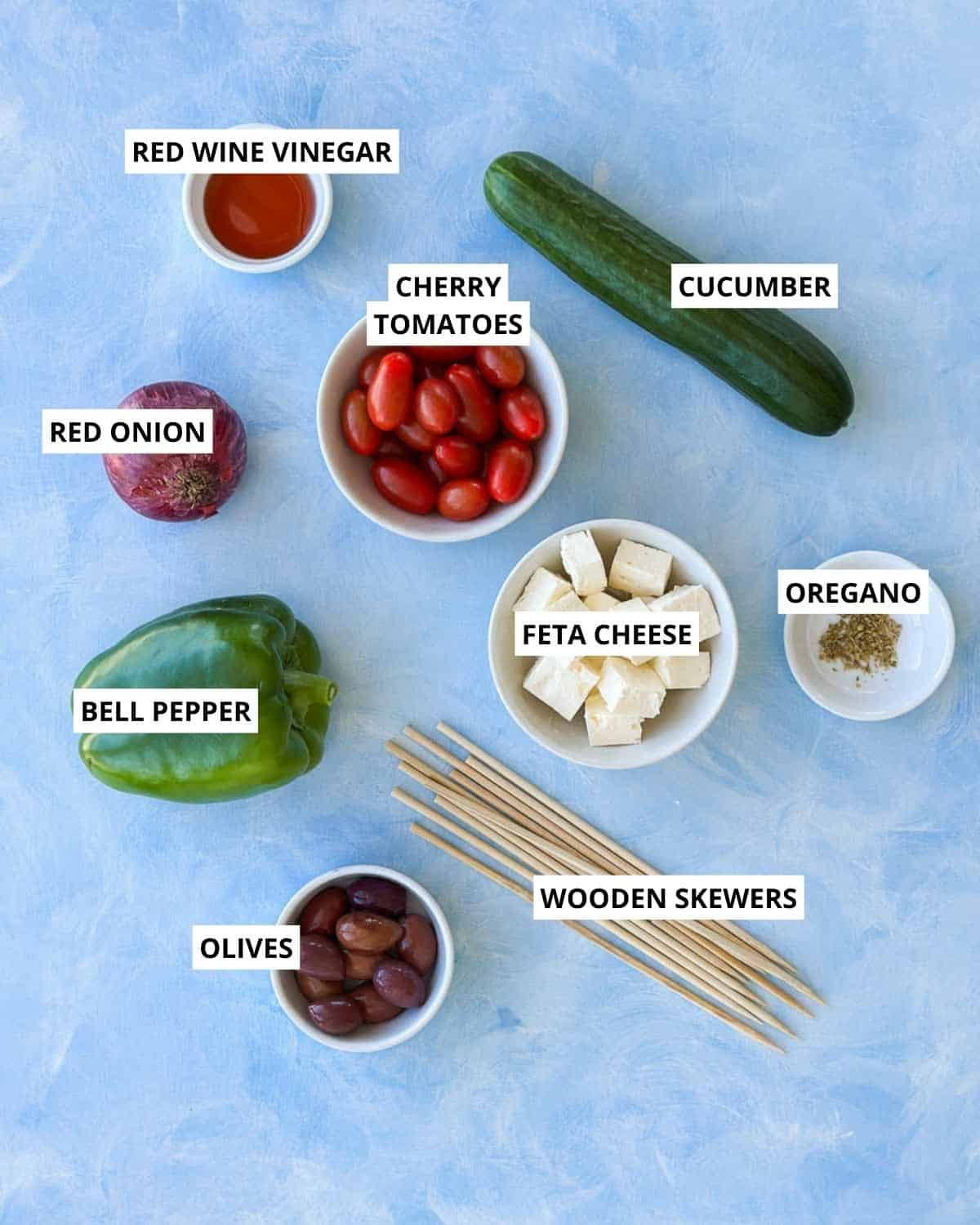 Ingredients for Greek salad skewers including cherry tomatoes, cucumber, feta cheese, Kalamata olives, red onion, bell pepper, oregano and red wine vinegar.