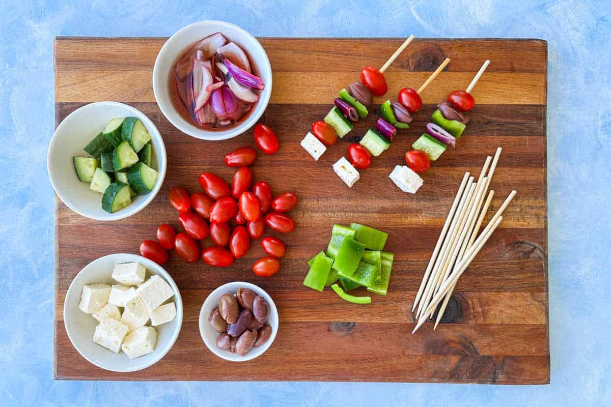 Greek salad skewer ingredients arranged on a wooden board with feta, cucumber, tomatoes, olives, pickled red onion and partially assembled skewers.