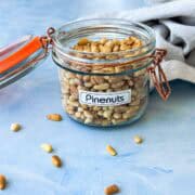 Glass jar filled with golden pine nuts on a blue surface with a soft linen cloth in the background