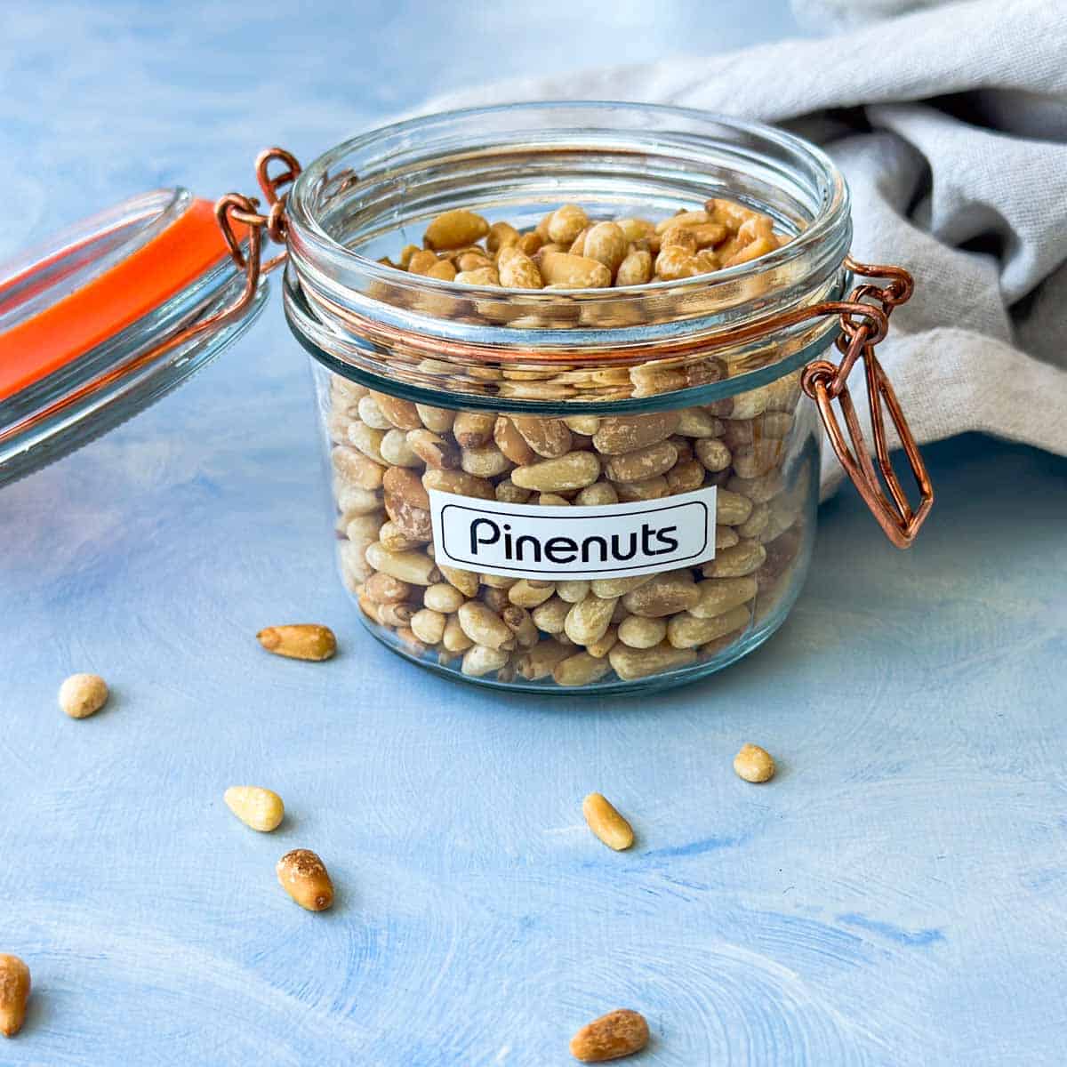 Glass jar filled with golden pine nuts on a blue surface with a soft linen cloth in the background