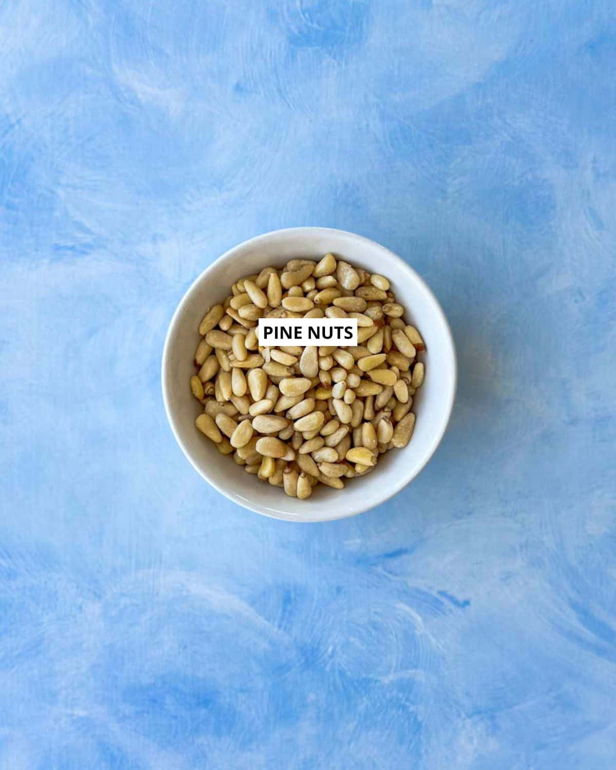 Overhead view of raw pine nuts in a white bowl on a blue background, labeled for the ingredient shot