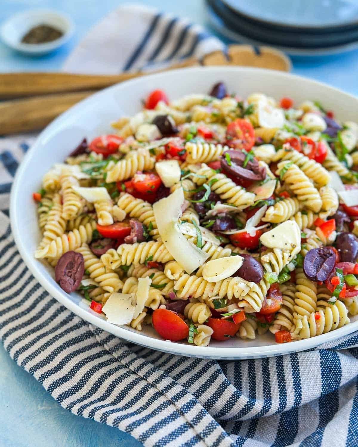 Angled view of Italian pasta salad in a shallow white bowl with rotini, olives, cherry tomatoes, fresh herbs and shaved parmesan.