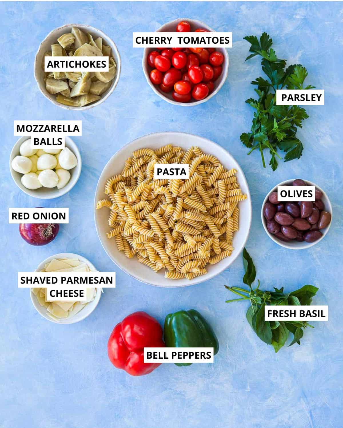 Ingredients for Italian pasta salad arranged on a blue surface including rotini pasta, cherry tomatoes, kalamata olives, mozzarella balls, artichokes, red onion, bell peppers, fresh basil, parsley and shaved parmesan.