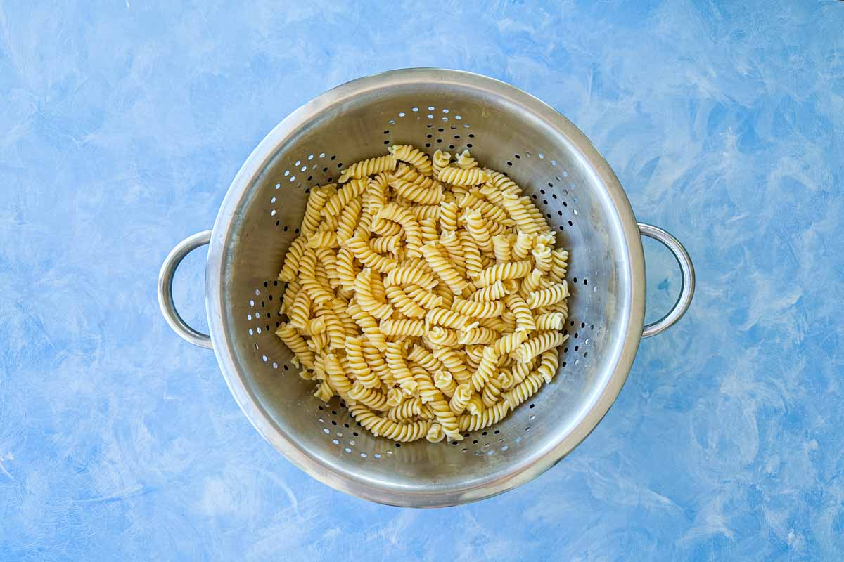 Top view of cooked rotini pasta draining in a metal colander on a blue surface for Italian pasta salad.