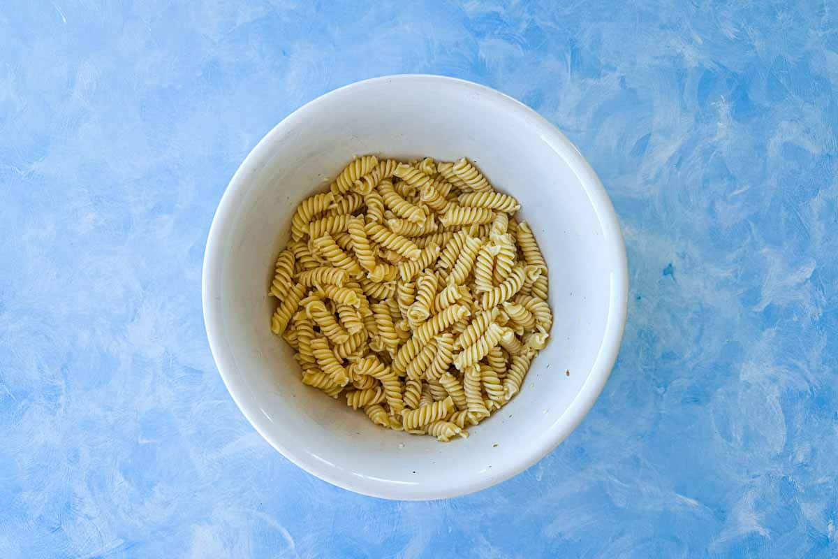 Top view of cooked rotini pasta in a large white mixing bowl on a blue surface ready to assemble Italian pasta salad.