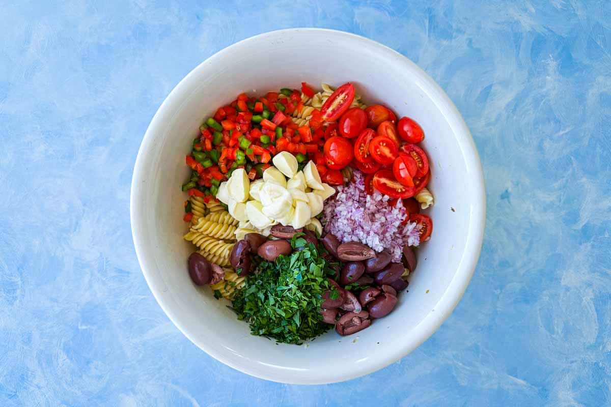 Top view of chopped vegetables, olives, mozzarella balls and fresh herbs added to pasta in a white bowl on a blue surface.