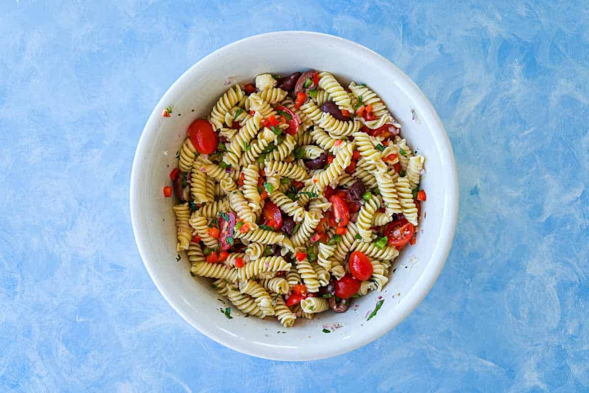Italian pasta salad being tossed with rotini, cherry tomatoes, olives, peppers, herbs and dressing in a white bowl.