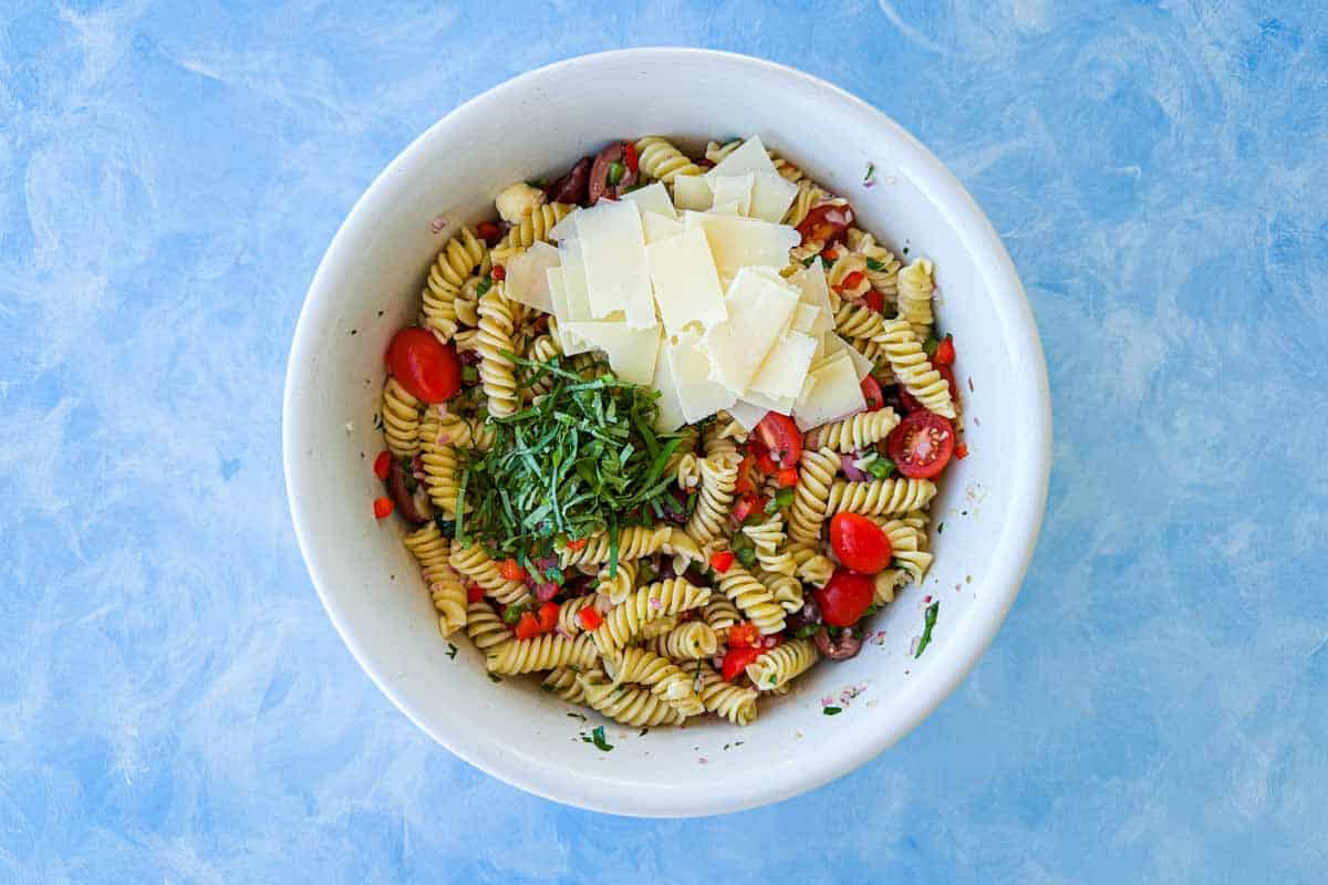 Italian pasta salad finished with fresh basil and shaved parmesan cheese in a large white mixing bowl on a blue surface.