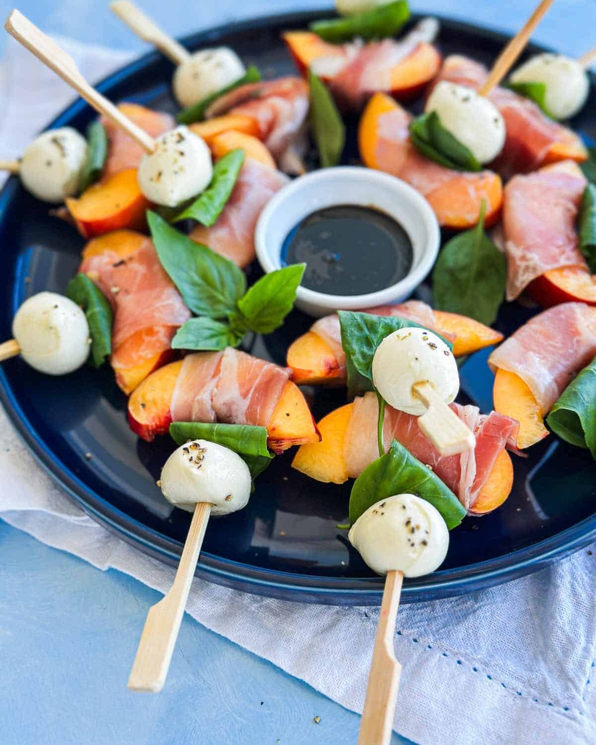 Close-up detail of peach wedges wrapped in parma ham with mozzarella balls and basil leaves on cocktail skewers, arranged on a dark plate with a small bowl of balsamic glaze in the center.