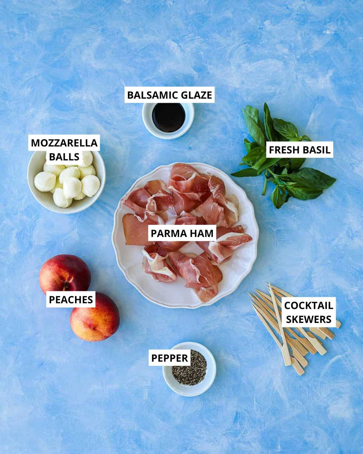 Overhead layout of ingredients including fresh peaches, parma ham, mozzarella balls, basil, balsamic glaze, pepper, and cocktail skewers arranged neatly on a light blue textured surface with labeled elements.