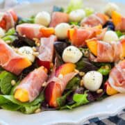 Close-up of peach and prosciutto salad with soft prosciutto, fresh peach slices, mozzarella balls, mixed leaves, and toasted pine nuts.