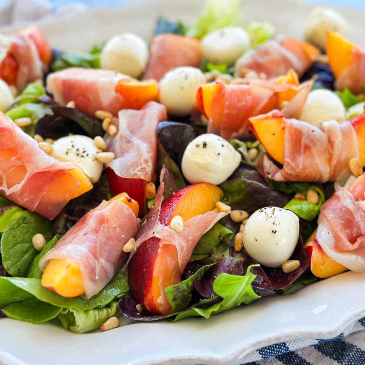 Close-up of peach and prosciutto salad with soft prosciutto, fresh peach slices, mozzarella balls, mixed leaves, and toasted pine nuts.