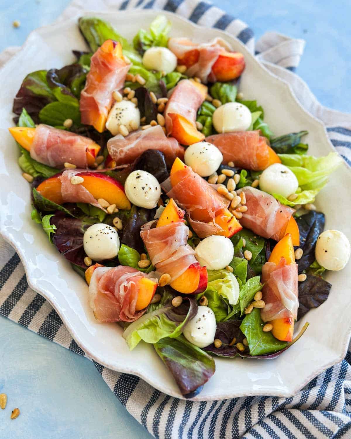Peach and prosciutto salad served on a white platter with mixed greens, basil, mozzarella balls, and toasted pine nuts.
