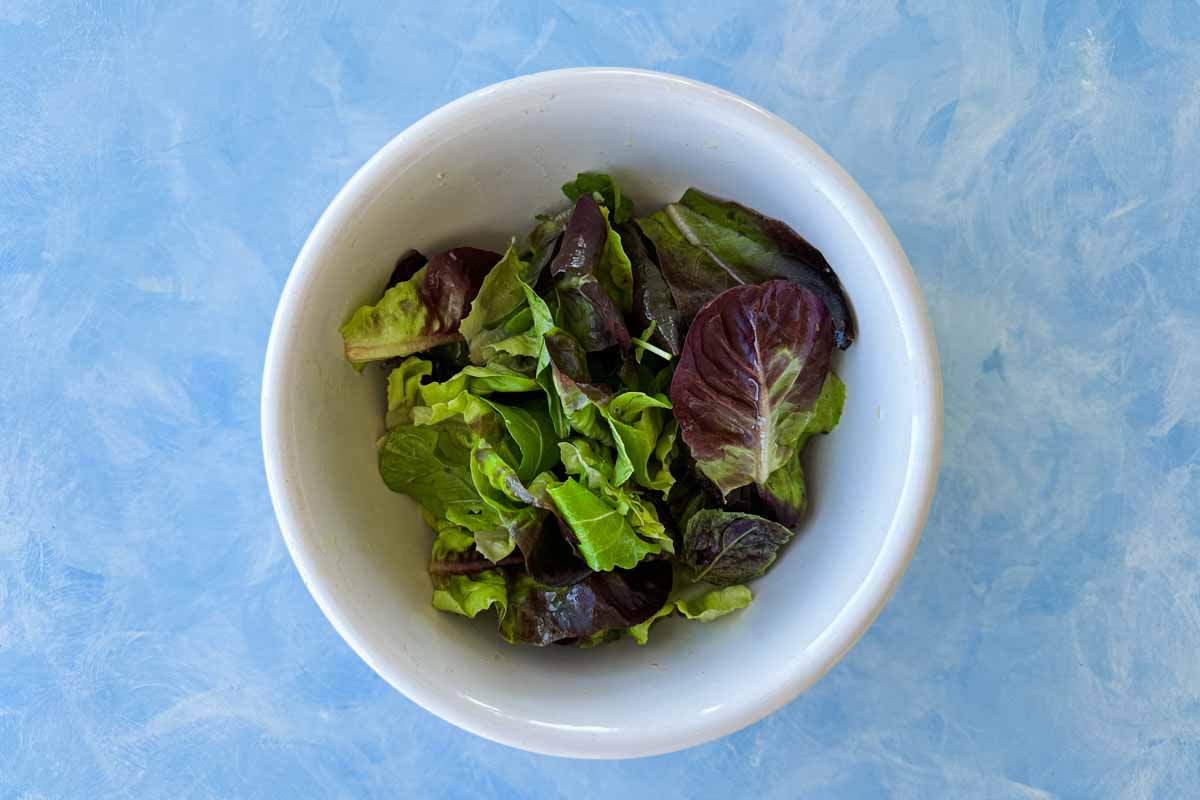 Mixed salad leaves in a white bowl on a blue surface, ready to form the base for peach and prosciutto salad.