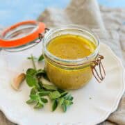 Close-up of a jar of red wine vinegar dressing with a rich golden color, surrounded by fresh herbs and garlic on a neutral linen setting.