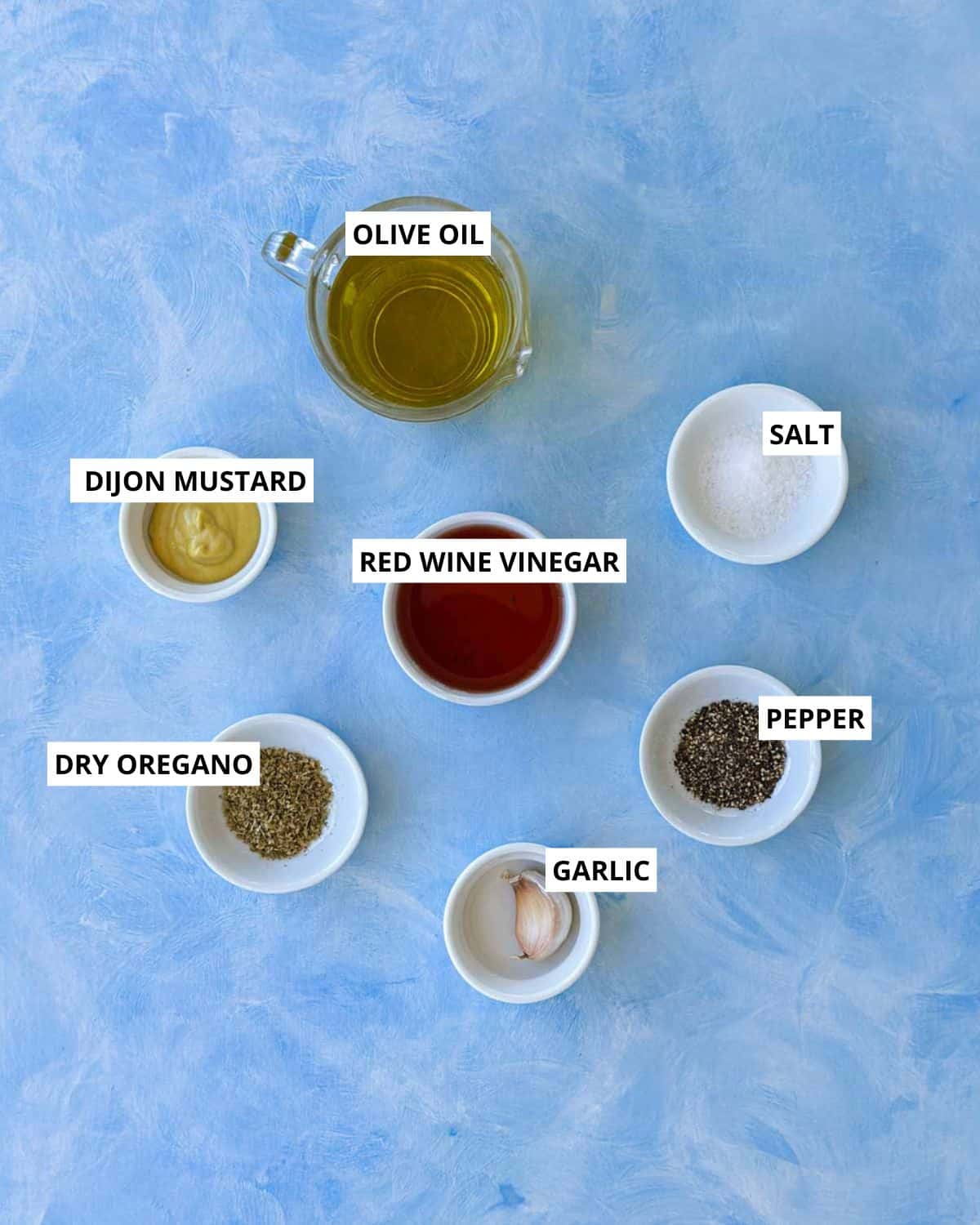 Flat lay of red wine vinegar dressing ingredients including olive oil, Dijon mustard, garlic, dried oregano, salt, pepper, and red wine vinegar on a soft blue background.