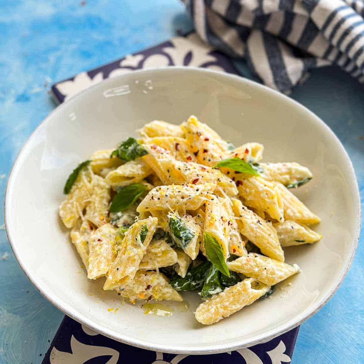 Creamy ricotta and lemon pasta plated in a white bowl with fresh basil leaves, parmesan cheese, and chili flakes on a light blue background.