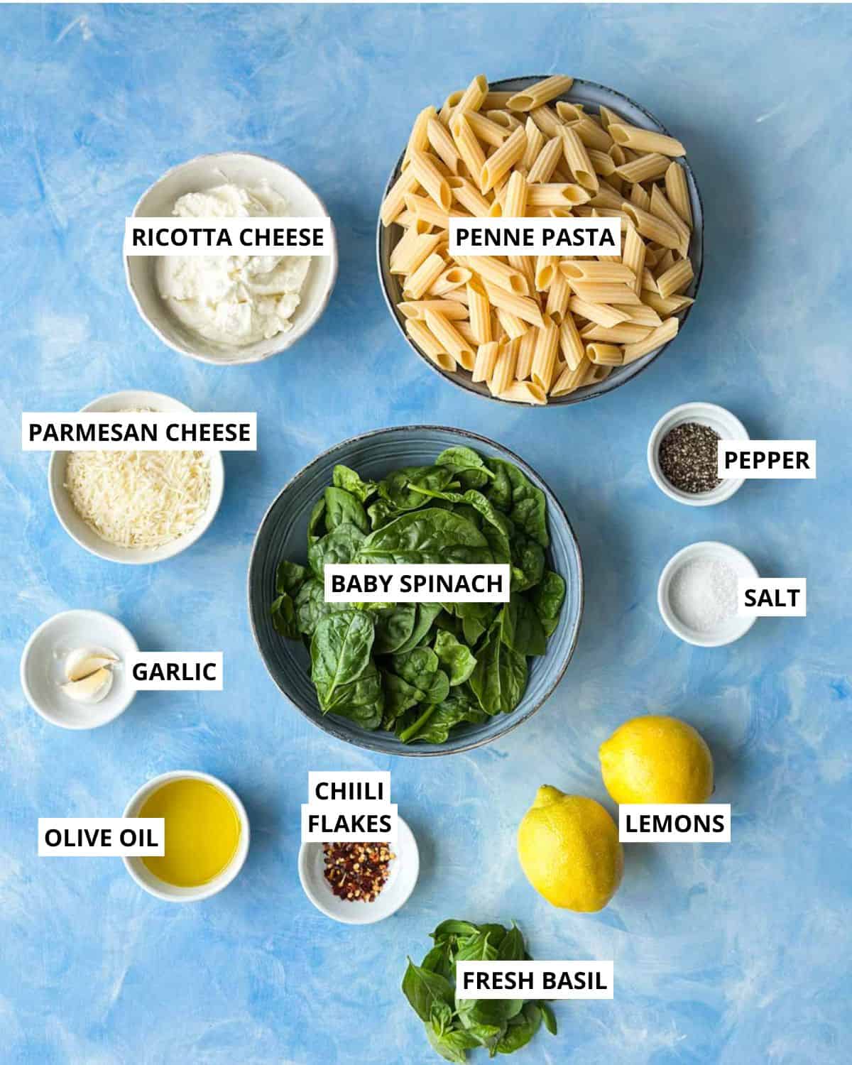 Flat lay of ricotta and lemon pasta ingredients including penne pasta, ricotta cheese, parmesan, baby spinach, garlic, olive oil, chili flakes, fresh basil, lemons, salt, and pepper on a blue background.