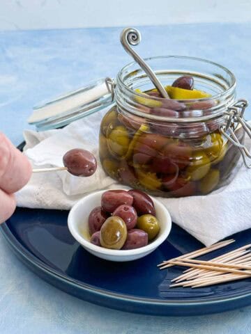 Side view of warm marinated olives in a glass jar with a hand holding a cocktail stick with an olive above a small serving bowl.