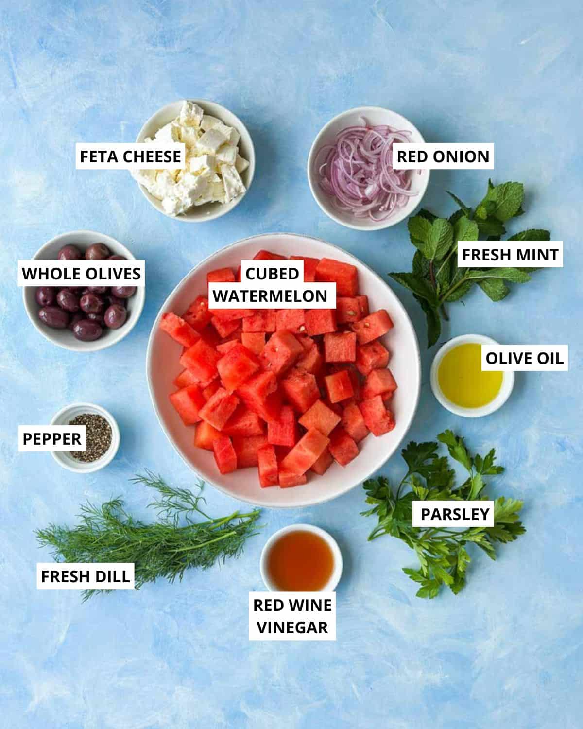 Flat lay of watermelon feta salad ingredients including cubed watermelon, feta cheese, red onion, olives, fresh herbs, olive oil, and red wine vinegar on a blue background.