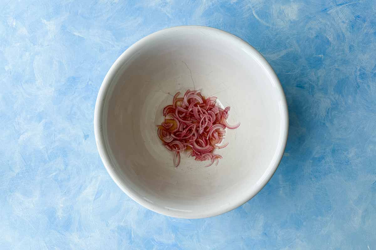 Thinly sliced red onion soaking in a bowl with vinegar, preparing a quick pickle for the watermelon feta salad