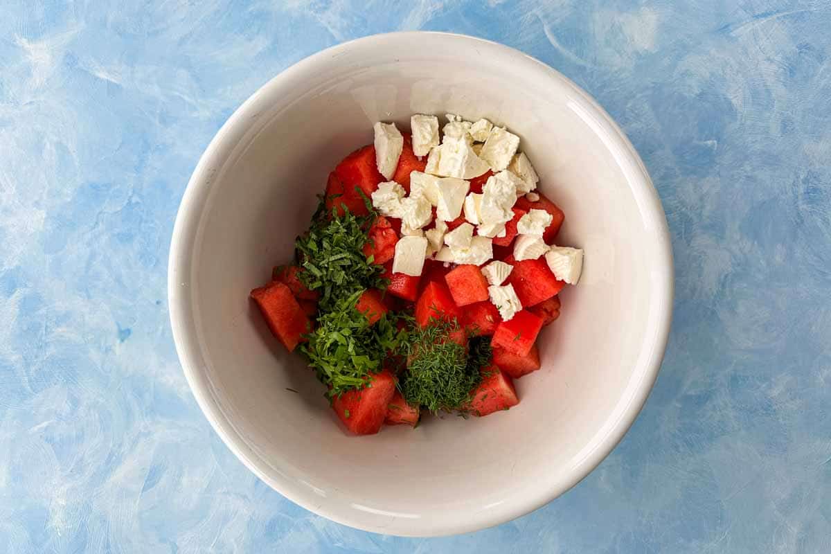 Cubed watermelon combined with crumbled feta, fresh herbs, and aromatics in a mixing bowl, ready to be tossed.