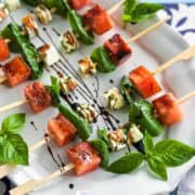 Close-up overhead view of watermelon feta skewers arranged neatly on a white serving plate, highlighting the contrast of sweet watermelon, herby feta cubes, and vibrant basil leaves with balsamic glaze.