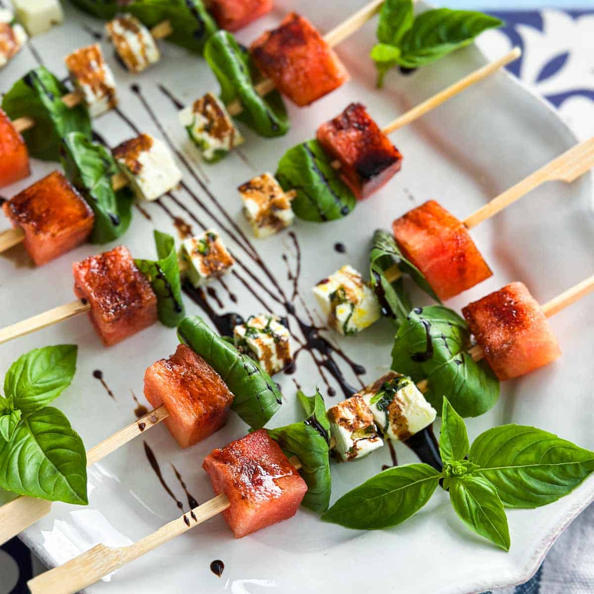 Close-up overhead view of watermelon feta skewers arranged neatly on a white serving plate, highlighting the contrast of sweet watermelon, herby feta cubes, and vibrant basil leaves with balsamic glaze.